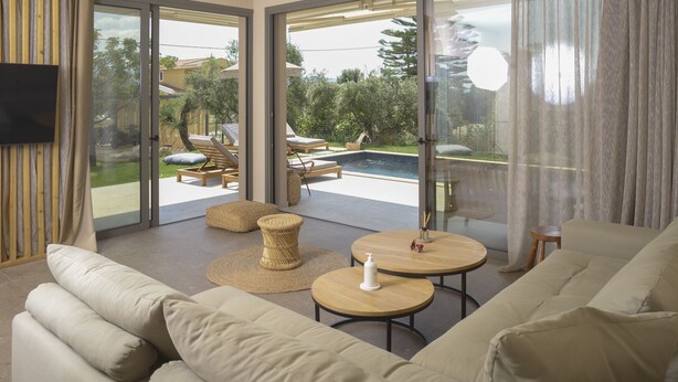 Un salon moderne avec un grand canapé, deux petites tables rondes en bois et une vue sur un jardin avec une piscine et des chaises longues.