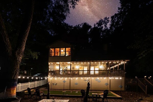 Une cabane en rondins à deux étages, décorée de guirlandes lumineuses, avec une terrasse et un petit espace porche.