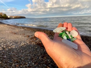 Une main tenant des petits galets de verre colorés sur une plage de galets, avec l’océan en arrière-plan.
