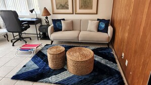 Una sala de estar con un sofá, dos cuadros enmarcados, un escritorio con una silla y un tapete azul y gris con dos cestas tejidas.