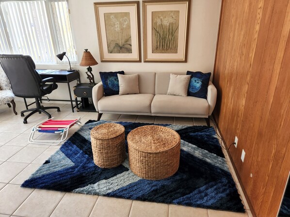 A living room with a sofa, two framed artworks, a desk with a chair, and a blue and gray rug with two woven baskets. - Oceanfront property with partial oceanview (Carlsbad)