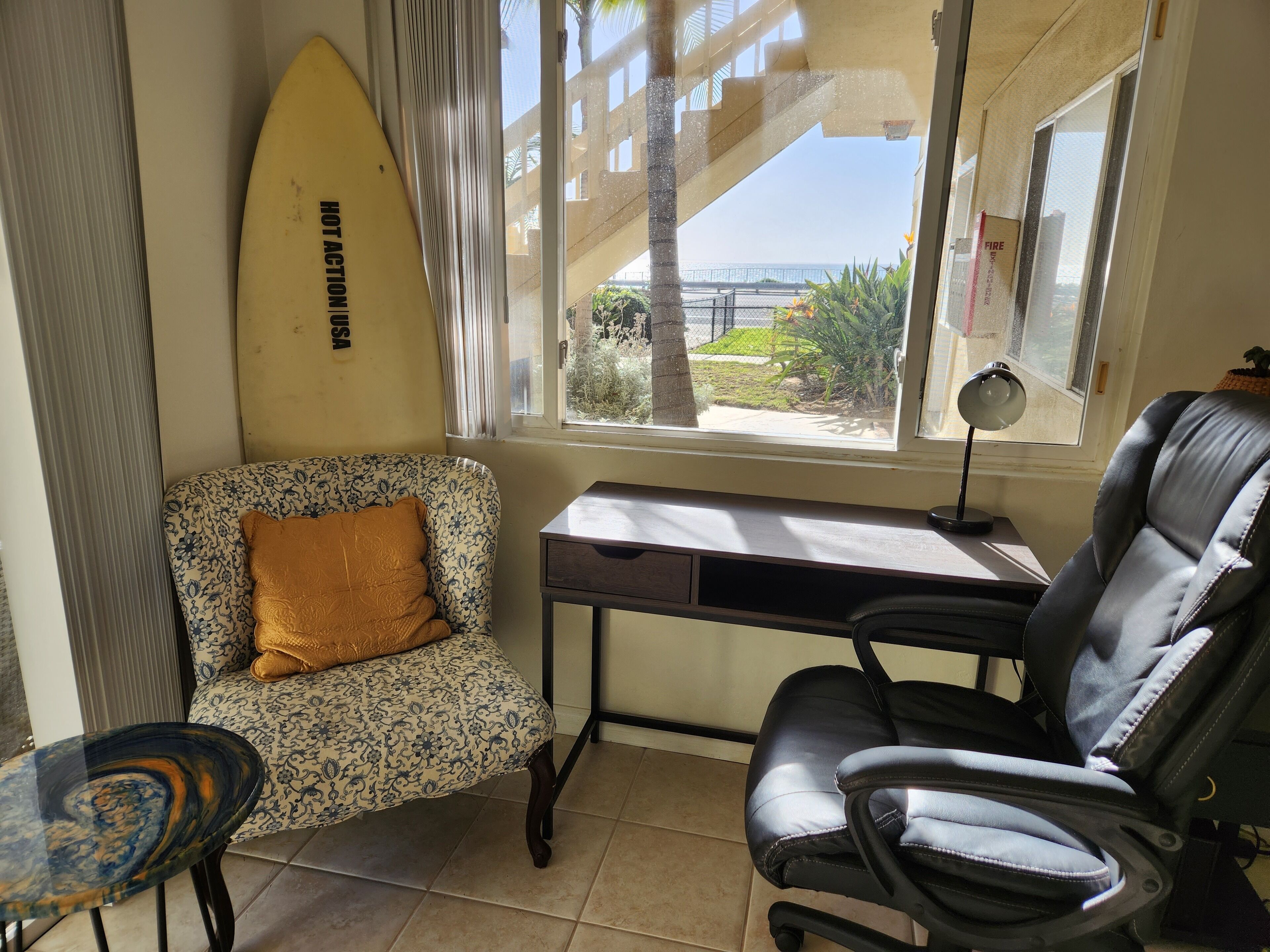Ein Zimmer mit Schreibtisch, Stuhl, Surfbrett und Blick ins Freie.