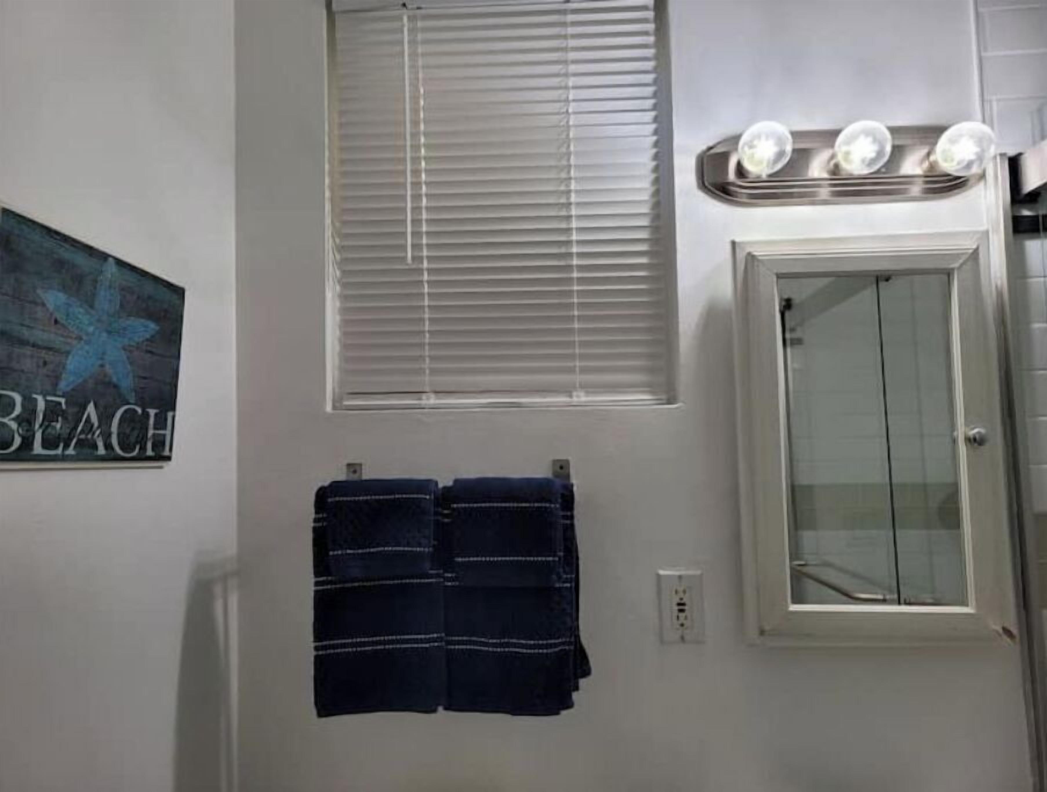 A bathroom with a towel rack holding two towels, a window with blinds, and a cabinet with lights above it.
