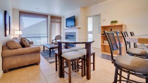 A modern living room with a sofa, a dining table with chairs, a TV mounted on the wall, and a bookshelf. - Resort-Style Condo w/ Lake Chelan & Mtn Views (Chelan)