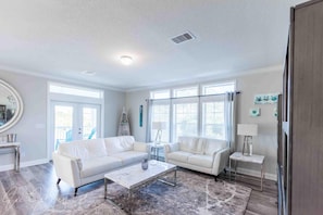 A spacious living room with a white sectional sofa, a coffee table, and a large window with blinds. - Hatchlife House - Steinhatchee (Dixie County)
