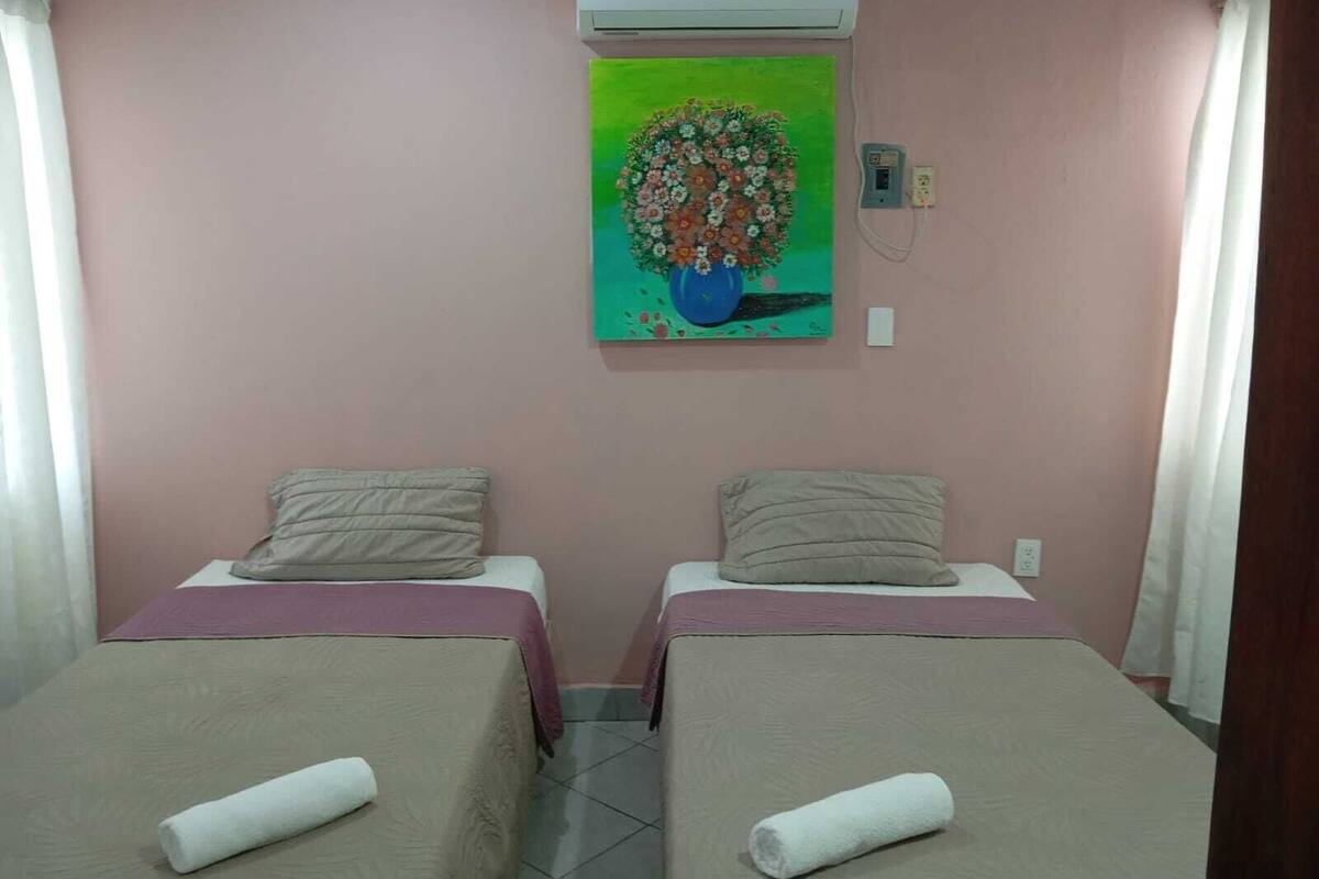A room with two beds, each with a pillow and a rolled towel. There is a painting on the wall above the beds.
