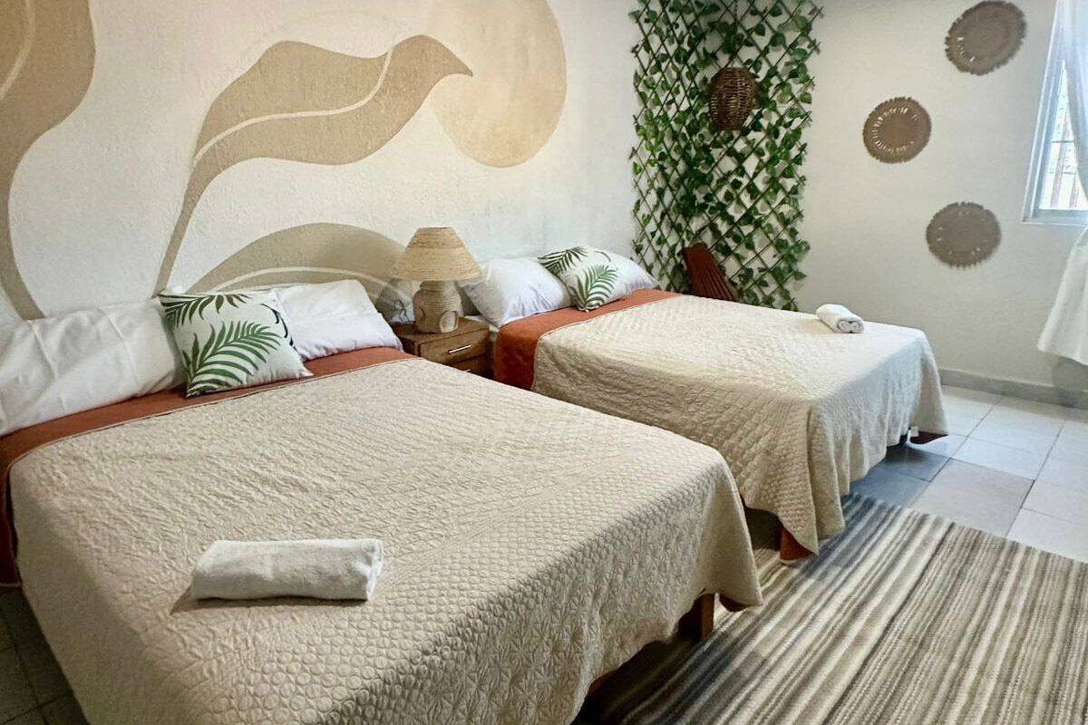 A room with two beds, each with a white bedspread and a pillow with a palm leaf pattern. There is a small table with a lamp between the beds, and a woven basket on the wall.