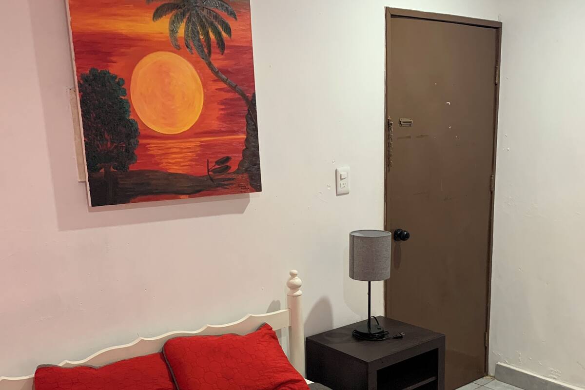 A bedroom with a bed, red pillows, a nightstand with a lamp, and a door.