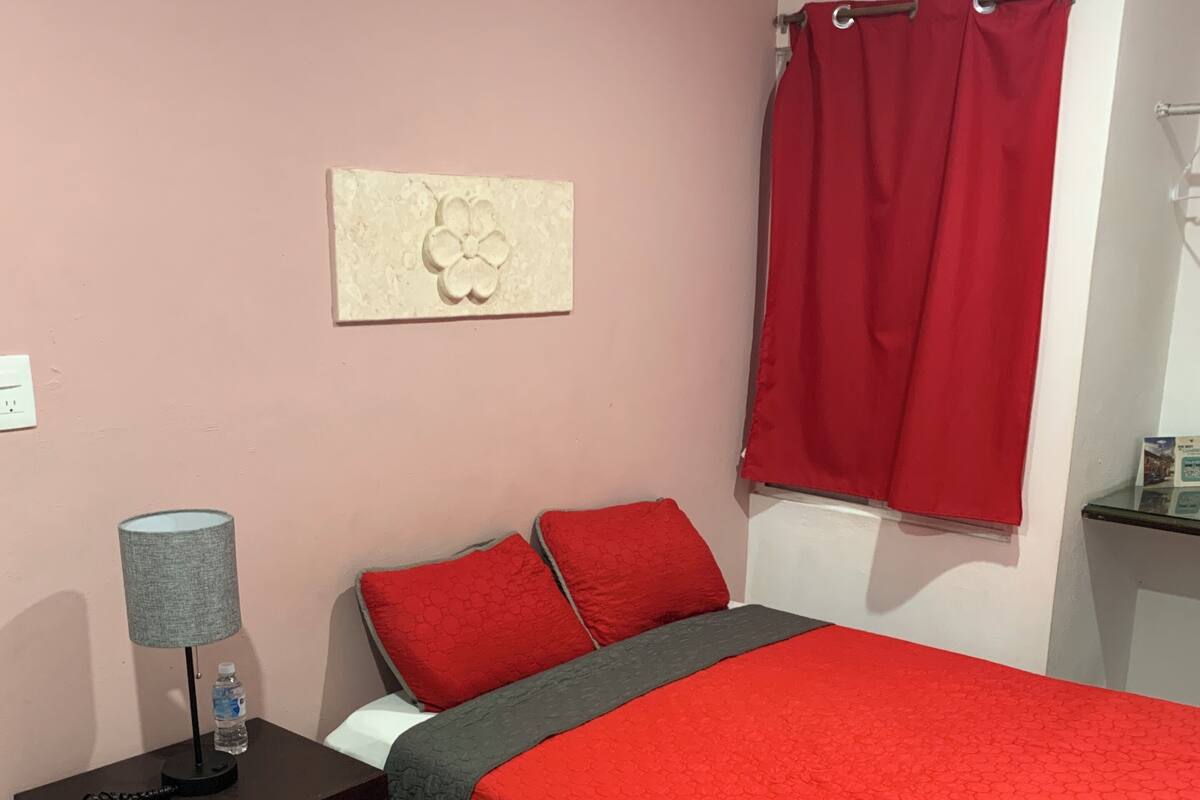 A bedroom with a red bedspread, two red pillows, a grey blanket, a grey lamp, a water bottle, and a wall decoration.