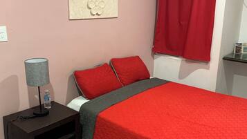 A bedroom with a red bedspread, two red pillows, a grey blanket, a grey lamp, a water bottle, and a wall decoration.