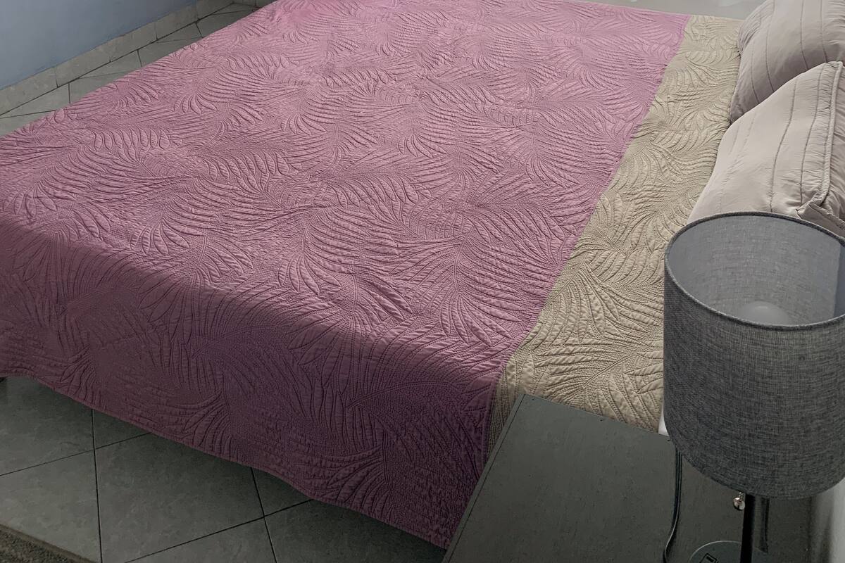 A bedroom with a pink textured bedspread, a bedside table with a lamp, a small shelf with bottles, and a window with curtains.