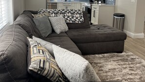 A modern living room with a sectional sofa and a kitchen in the background. - Cozy House with Large Deck (Hudson)