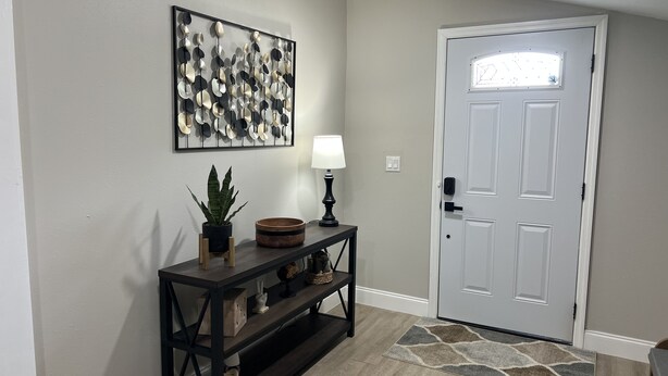 A hallway with a wooden console table, a potted plant, a wall-mounted abstract art piece, a door, and a patterned rug. - Cozy House with Large Deck (Hudson)
