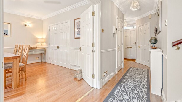 A spacious hallway with wooden flooring, white doors, and a patterned rug leading to a dining area with wooden chairs and a table.