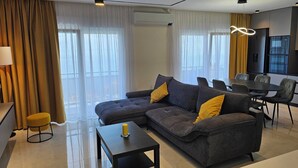 Ein modernes Wohnzimmer mit einem grauen Sofa, gelben Kissen, einem kleinen Tisch und einem Essbereich mit Stühlen.