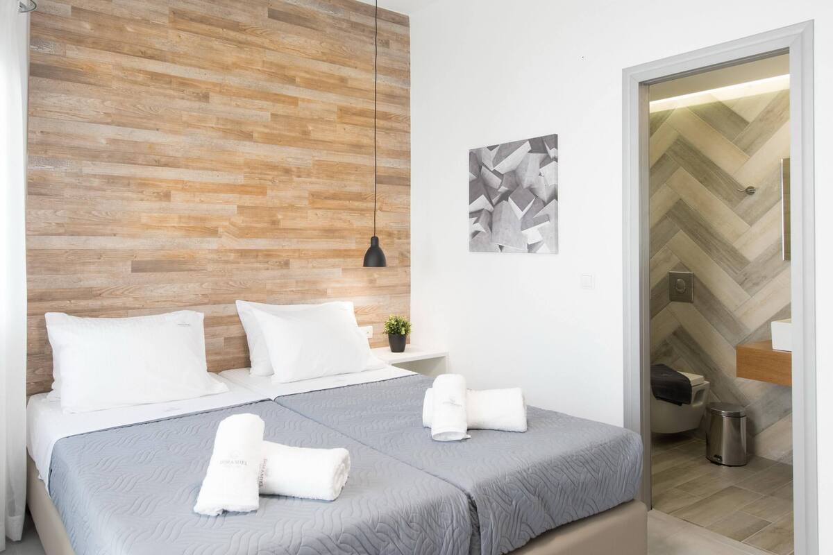 A modern hotel room with a wooden headboard, a grey quilted bedspread, two white pillows, and neatly folded towels on the bed.