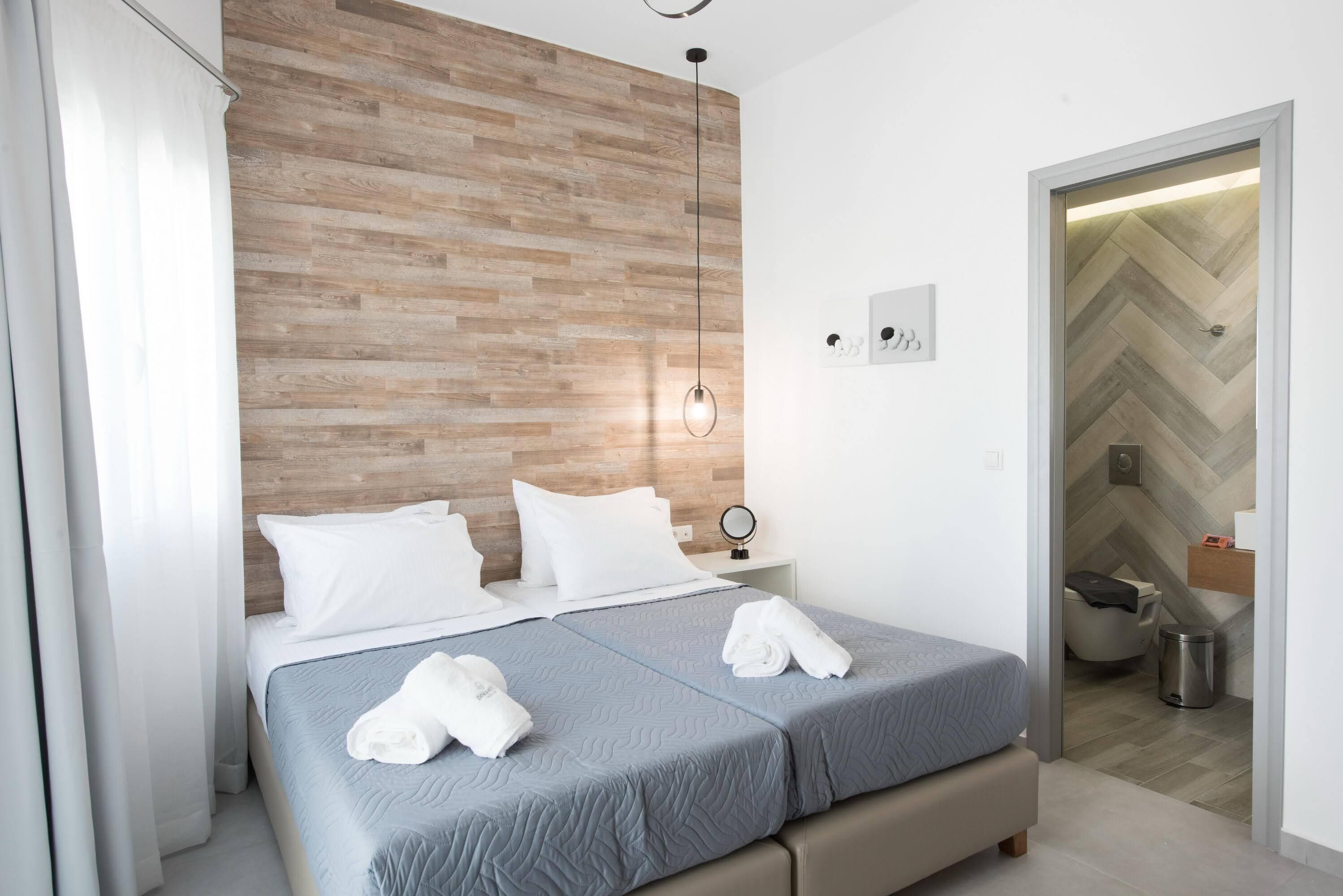 A modern hotel room with a wooden headboard, a bed with two pillows, a bedside table with a lamp, and a view of the bathroom through an open door.