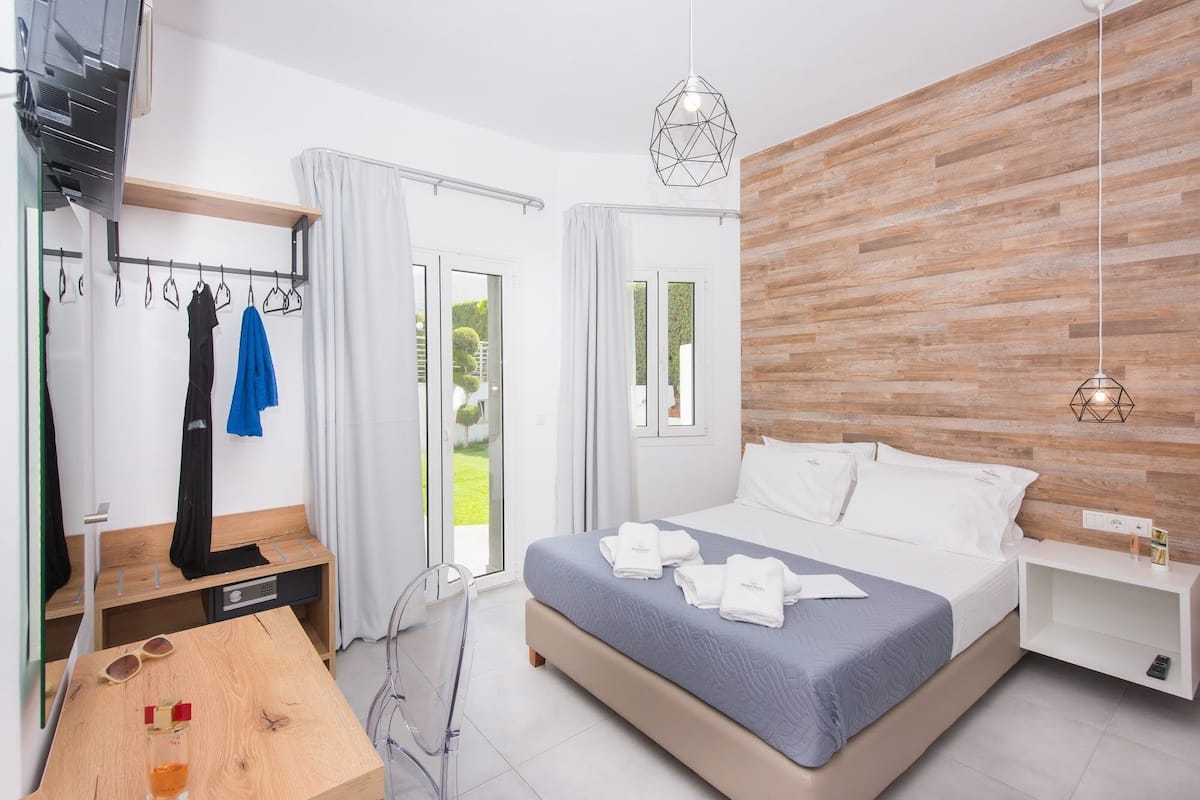 A modern hotel room with a bed, a wooden accent wall, a hanging light fixture, and a view of the outdoors.