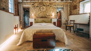 Un dormitorio rústico con vigas de madera a la vista, una pared de piedra, una cama con colcha acolchada, un cofre de madera y un escritorio con silla.