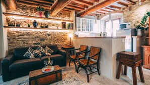 Una sala de estar rústica con pared de piedra, vigas de madera, un sofá, sillas, una mesa de centro con una botella y vasos, y una zona de cocina con microondas y refrigerador.