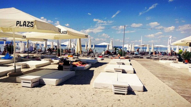 A beachside lounge area with multiple white lounge chairs and sunbeds, shaded by white umbrellas labeled "PLAZS". - Holiday apartment near the beach with internet (Siófok)