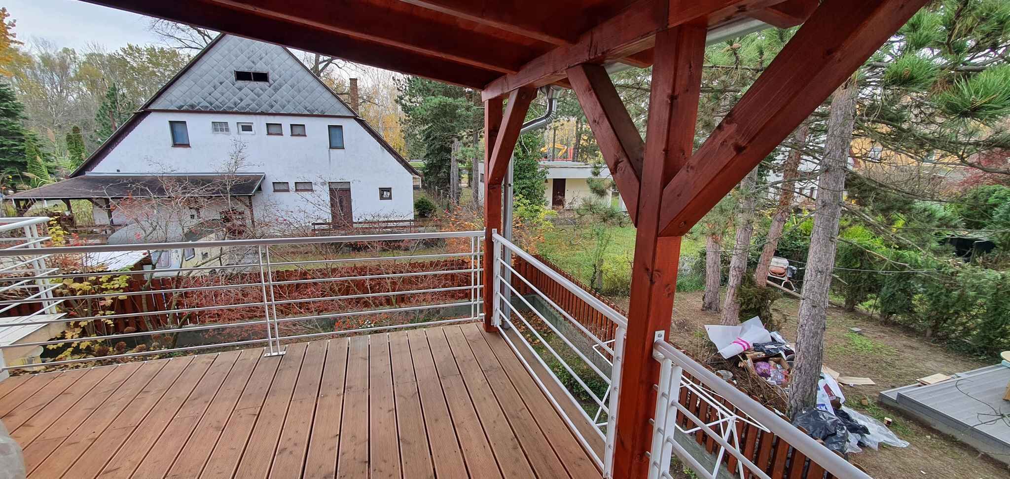 A wooden deck with a metal railing, overlooking a white house with a steep roof and a small attached structure.