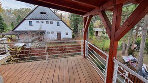 Eine Holzdar Terrasse mit Metallgeländer, mit Blick auf ein weißes Haus mit steilem Dach und einem kleinen Anbau.