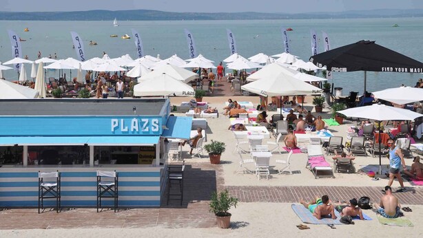 Ein Strandabschnitt mit mehreren Sonnenschirmen, Liegestühlen und einem Schild mit der Aufschrift 'PLAZA'.