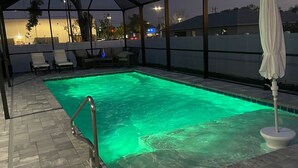 Una piscina exterior techada con fuente iluminada en verde, sillas de descanso y una sombrilla blanca.