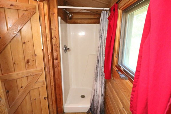 Une cabane en bois avec une cabine de douche, une fenêtre ornée d’un rideau rouge et une porte au motif en bois.