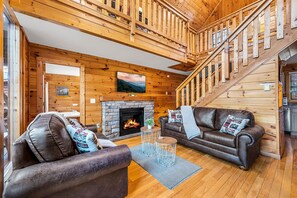 A cozy log cabin interior with a fireplace, leather sofas, and a wooden staircase.
