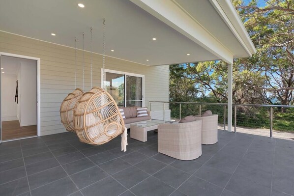 A covered outdoor area with a wicker swing chair, a sofa, and a dining table.