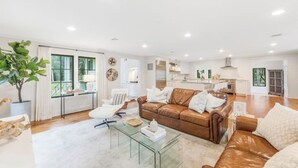 Un salon spacieux avec un canapé en cuir marron, une table basse en verre et un fauteuil blanc. Une grande fenêtre laisse entrer la lumière naturelle et offre une vue sur l’extérieur.