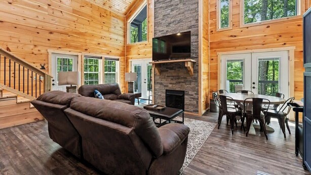 A spacious living room with a fireplace, wooden walls, and large windows. - 3BR 2BA w/ King Bed/Hot Tub in Heart of RRG (Campton)