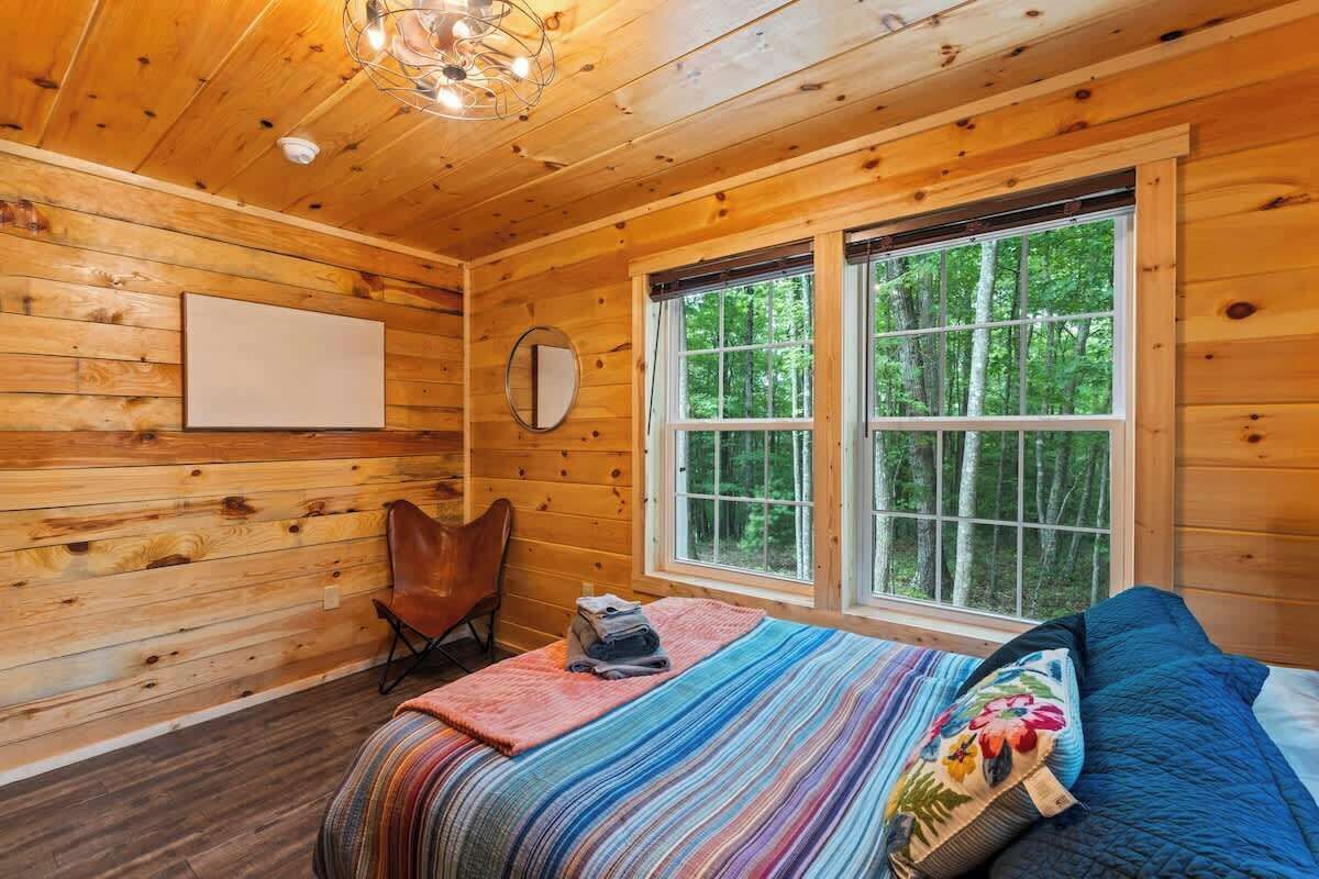 Ein gemütliches Blockhauskabinenzimmer mit Holzwänden, einem großen Fenster, einem Bett mit bunter Decke und einem Sessel.