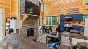 A cozy cabin interior with a stone fireplace, a flat-screen TV, a dining area, and a kitchen with blue cabinets. - 3BR 2BA w/ King Bed/Hot Tub in Heart of RRG (Campton)