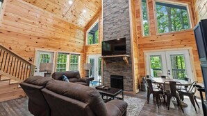 A spacious cabin interior with a high ceiling, wooden walls, a stone fireplace, a flat-screen TV, a dining area, and large windows offering views of the surrounding forest. - 3BR 2BA w/ King Bed/Hot Tub in Heart of RRG (Campton)