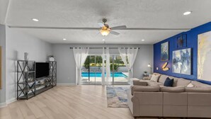 A spacious living room with a beige sofa, a TV on a metal stand, a ceiling fan, and a view of a swimming pool through large windows. - Luxury Ranch 2 Master Kg Bed w/ heated Salted Pool (Orlando)