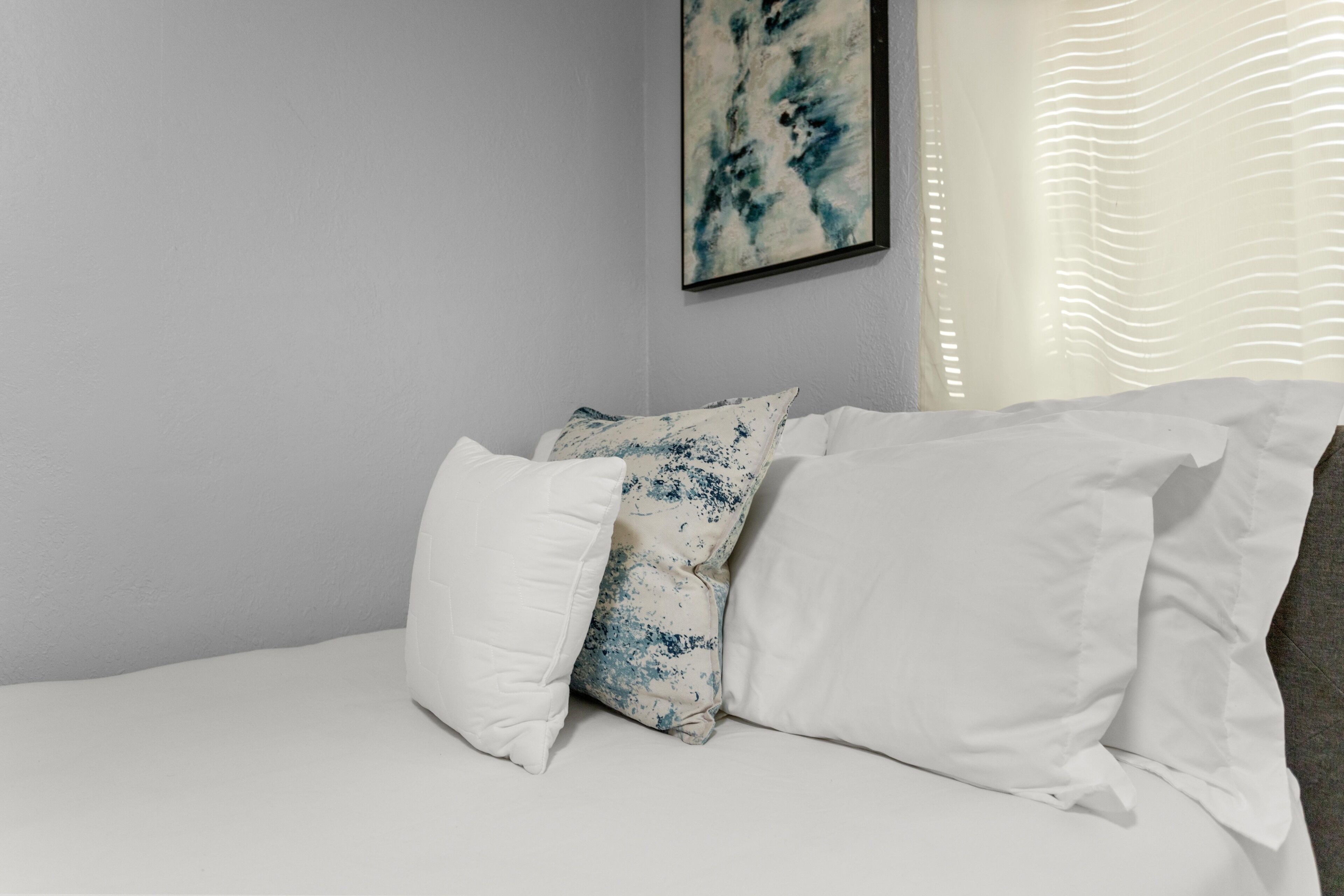 A neatly made bed with white and patterned pillows, a framed abstract painting on the wall, and a window with blinds.