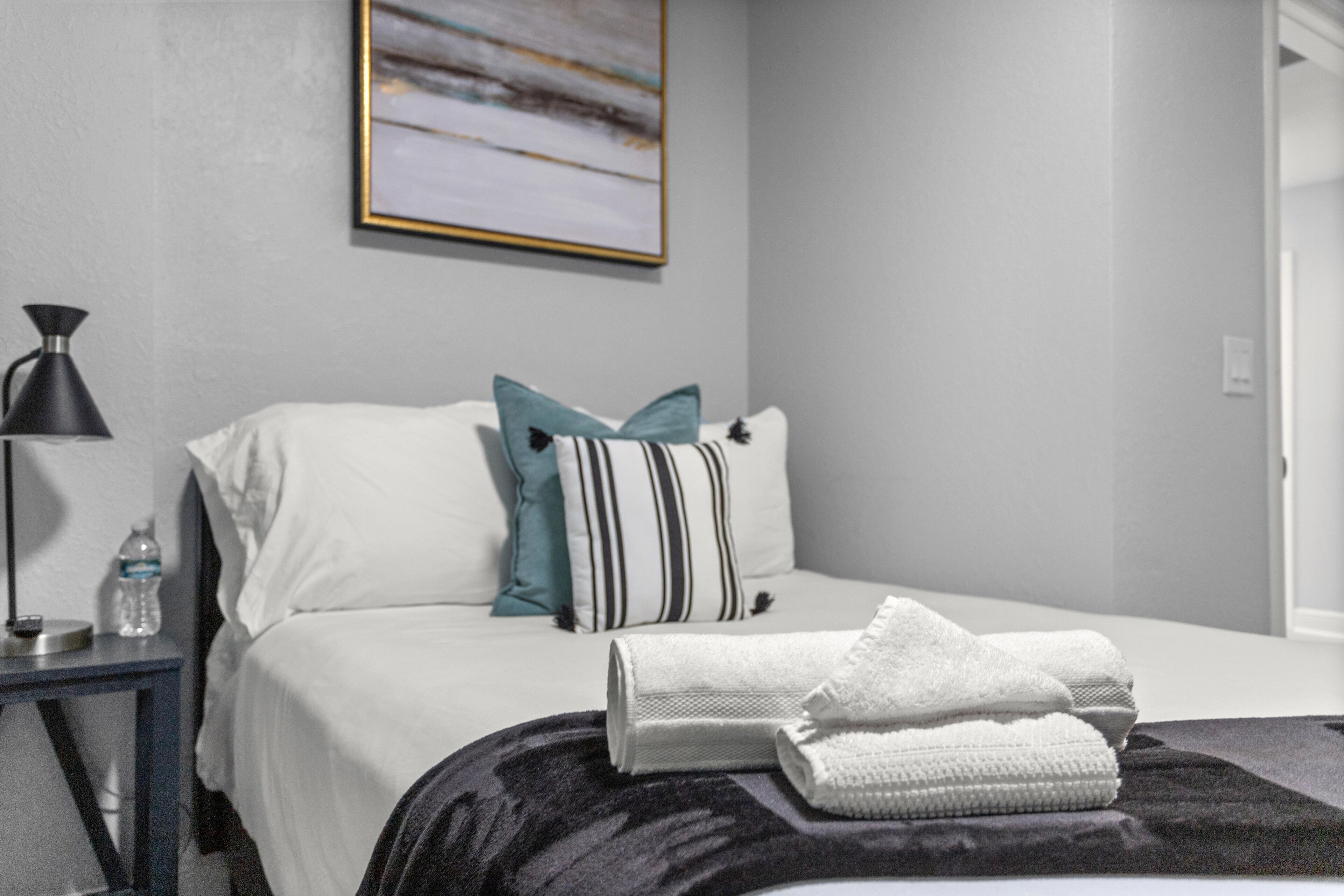 Een net opgemaakt bed met witte en gestreepte kussens, een zwarte lamp en een waterfles op een nachtkastje.