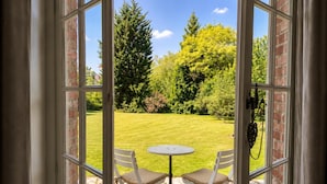 A view through a glass door to a patio with a table and two chairs, a brick wall, and a green lawn with trees. - les jardins de Beaumont (Hem)