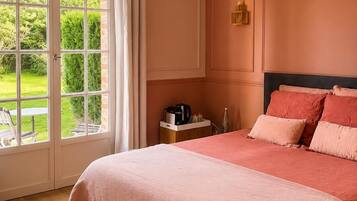 A bedroom with a large window, a bed with pink bedding, a nightstand with a coffee maker, and a view of a garden.