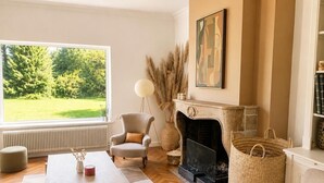 A living room with a fireplace, a large window with a view of greenery, a round floor lamp, and a bookshelf. - les jardins de Beaumont (Hem)