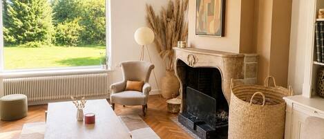 A living room with a fireplace, a large window with a view of greenery, a round floor lamp, and a bookshelf.