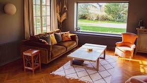 A living room with a sofa, coffee table, and armchair, a large window with a view of a garden, and a radiator. - les jardins de Beaumont (Hem)