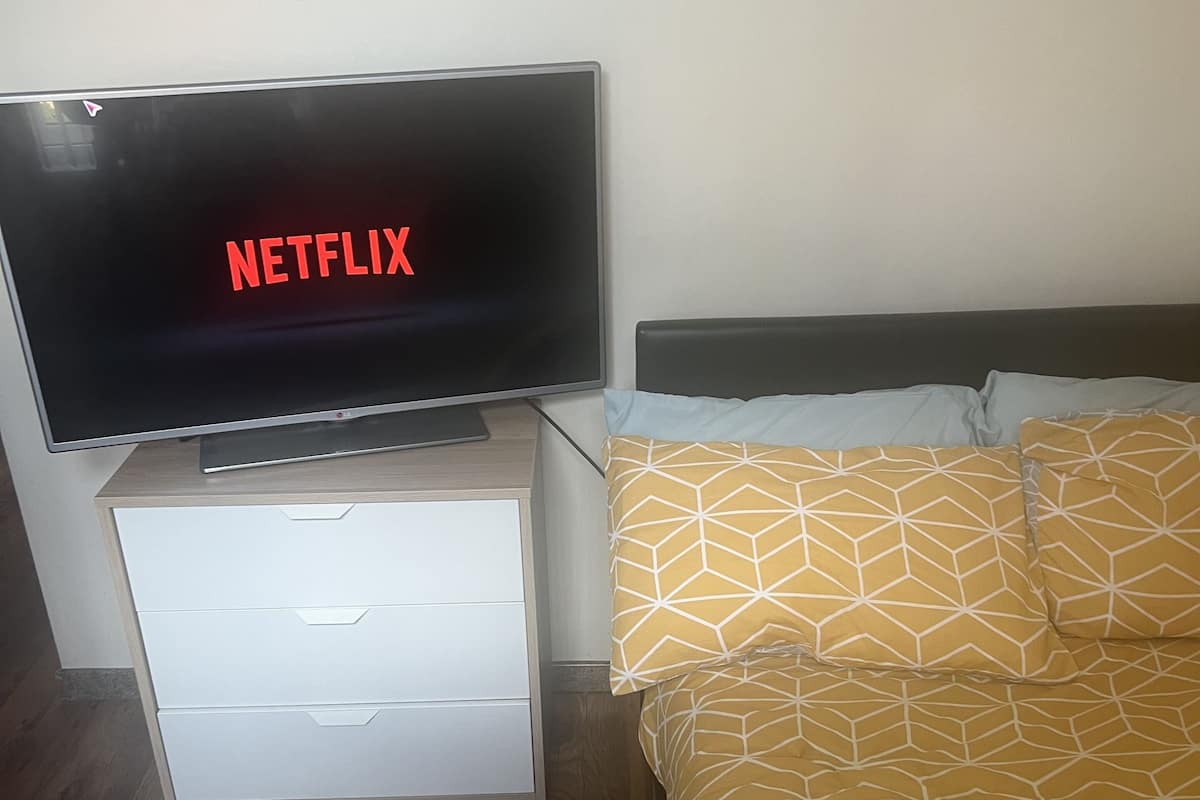 A room with a television displaying 'NETFLIX' in red letters, a white bedside table with drawers, a bed with patterned bedding, and a wooden floor.