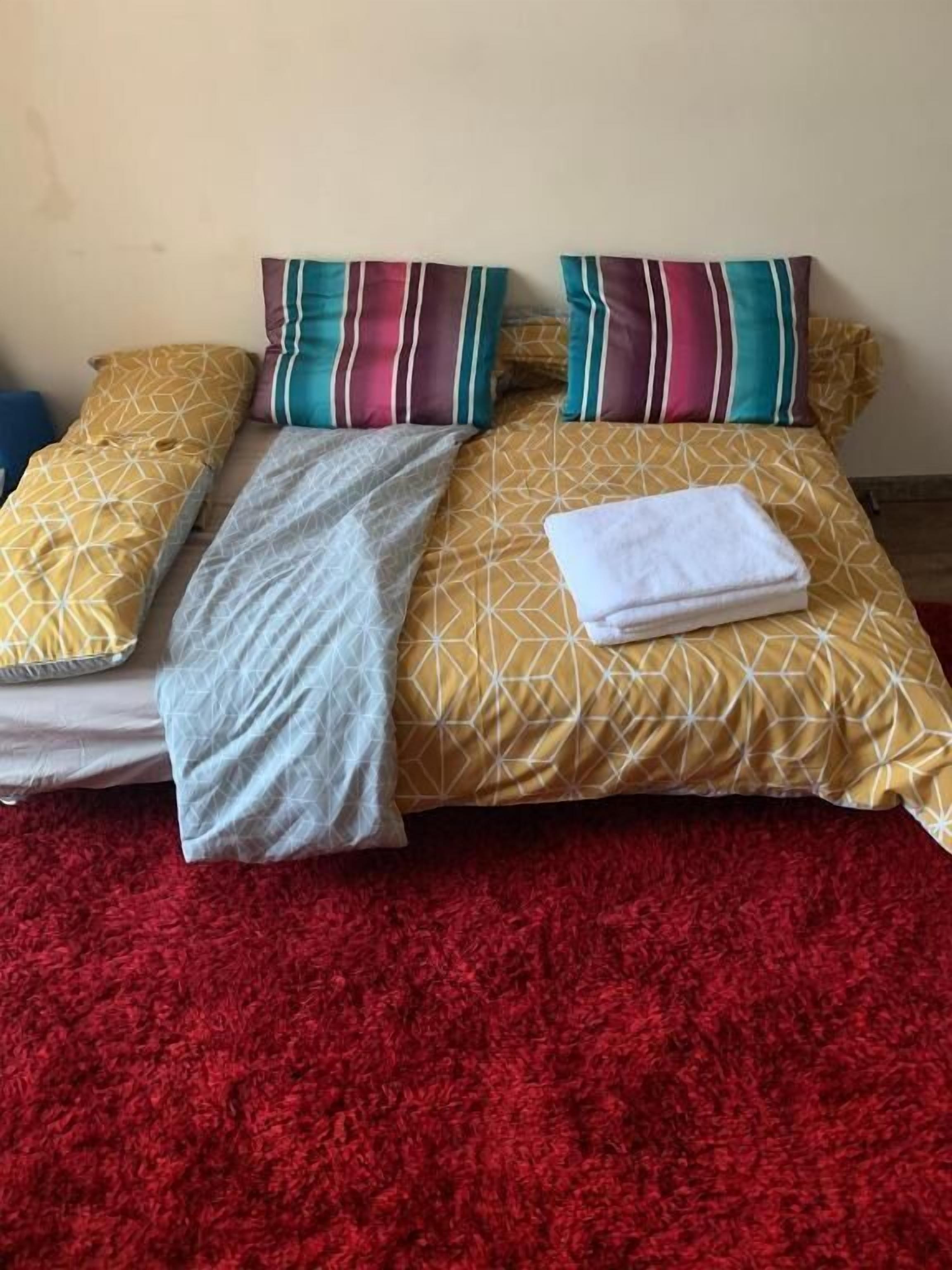 Een bed met beddengoed met een patroon en kussens, een opgevouwen witte handdoek en een rood vloerkleed.