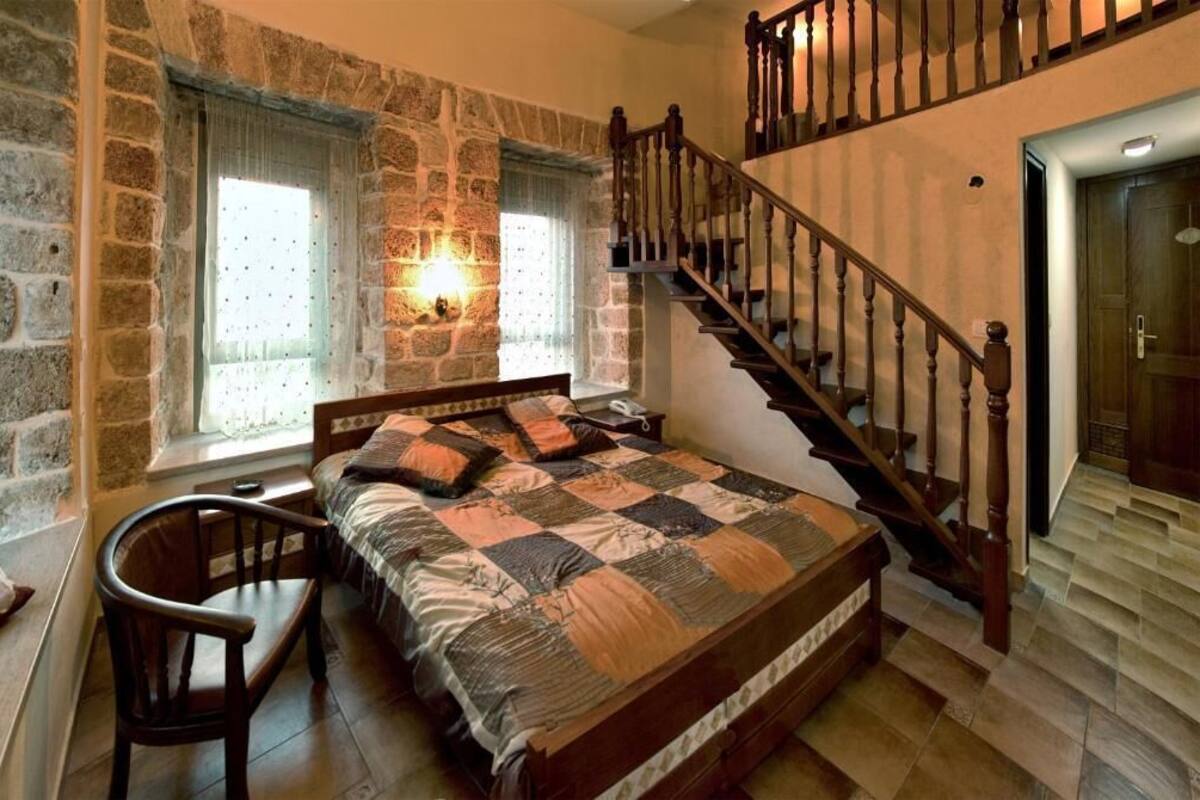 Een slaapkamer met een stenen muur, een houten bed, een stoel, een raam met gordijnen en een trap.