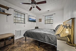 Quarto com cama, um banquinho, ventoinha no teto e duas janelas com persianas.