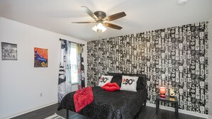 A bedroom with a ceiling fan, a bed with black bedding and red pillows, a patterned wallpaper, and a small table with a lamp. - 90's in Nash! 2bd Townhome in WEST END! 8 min ->DT! (Nashville)